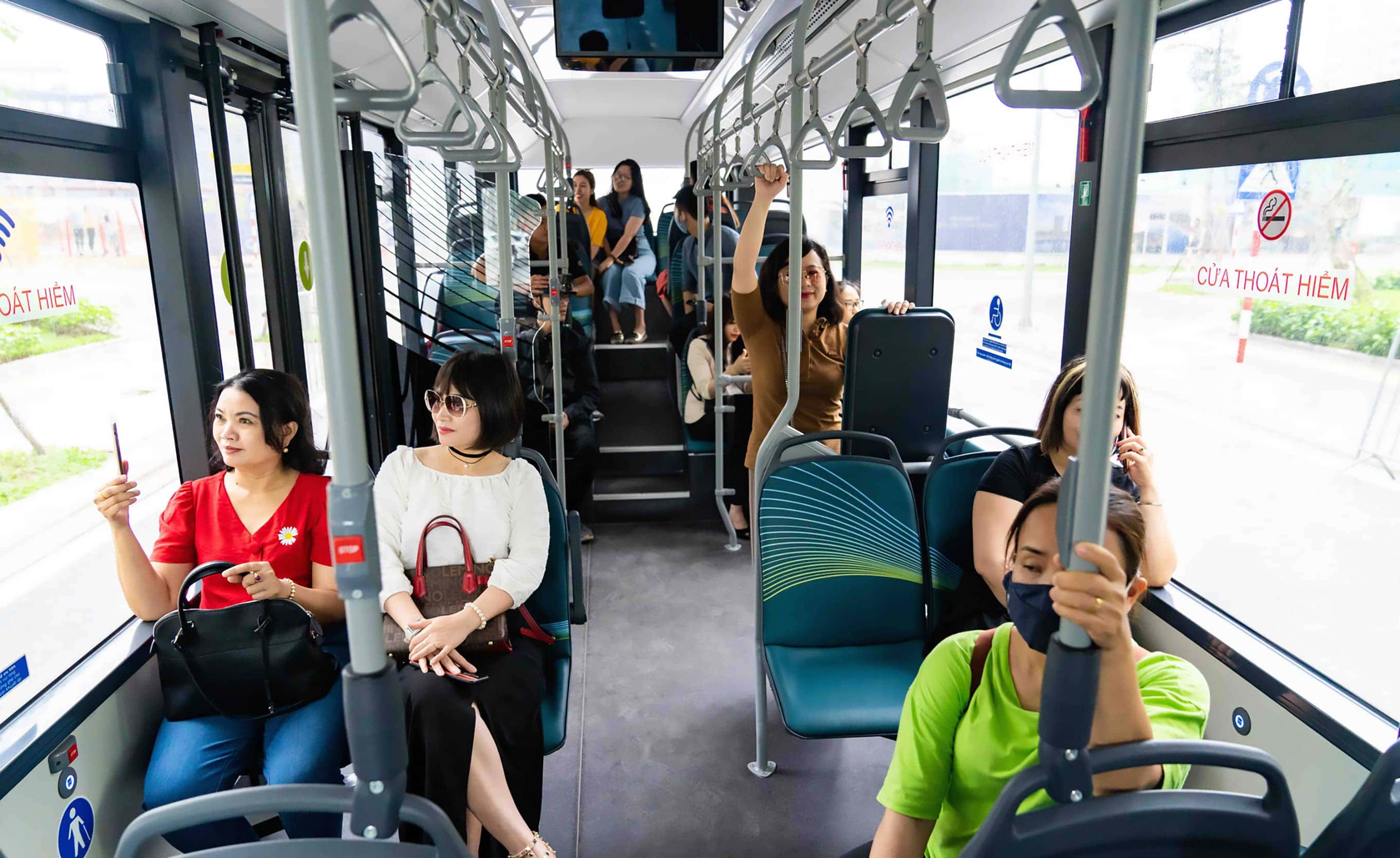 Một nhóm khách du lịch hoặc cư dân đang đứng đợi xe bus tại một điểm dừng xe trong khu đô thị Vinhomes Ocean Park 1, phía sau là hàng cây xanh và các tòa nhà