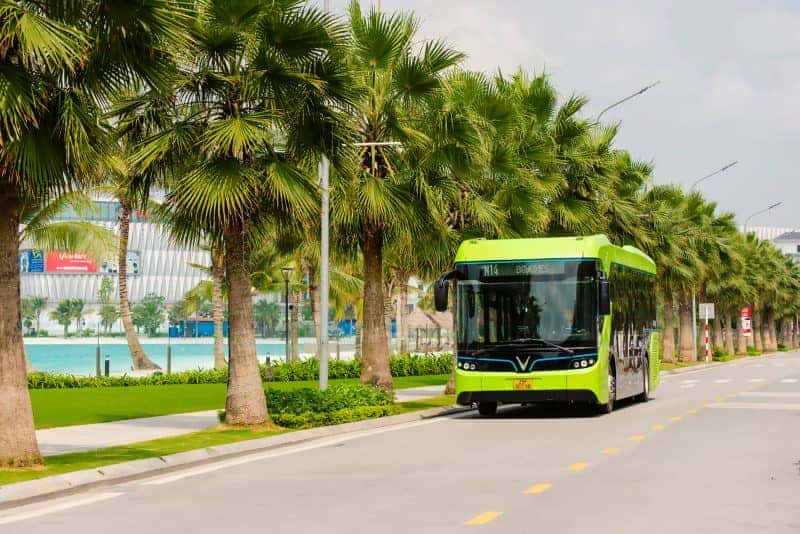 Xe bus điện VinBus màu xanh và đen đang chạy trên đường trong khuôn viên khu đô thị Vinhomes Ocean Park 1 với các tòa nhà cao tầng và cây xanh